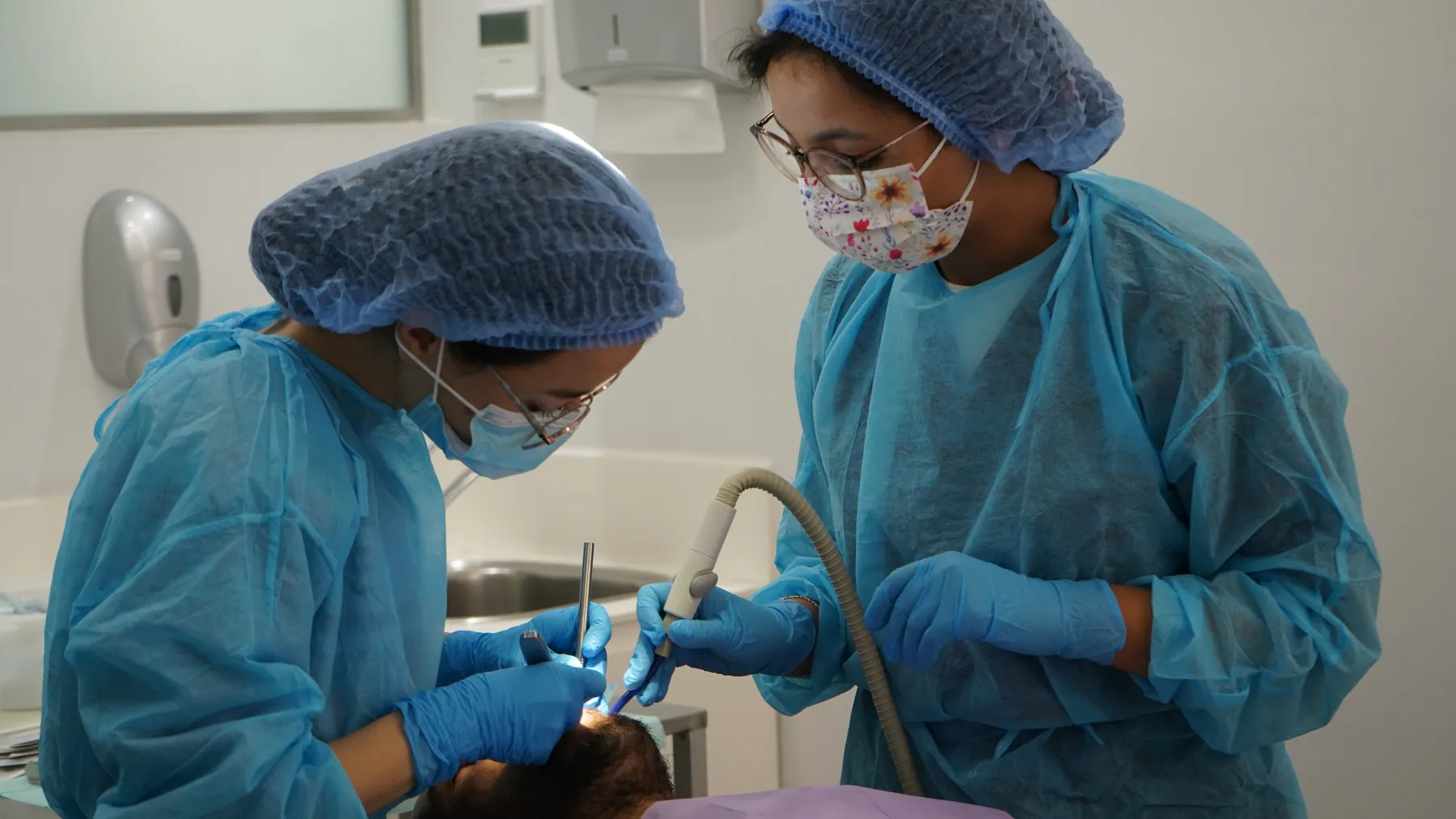Deux praticiennes en tenue chirurgicale opèrent, instrument et aspiration en main, sous supervision